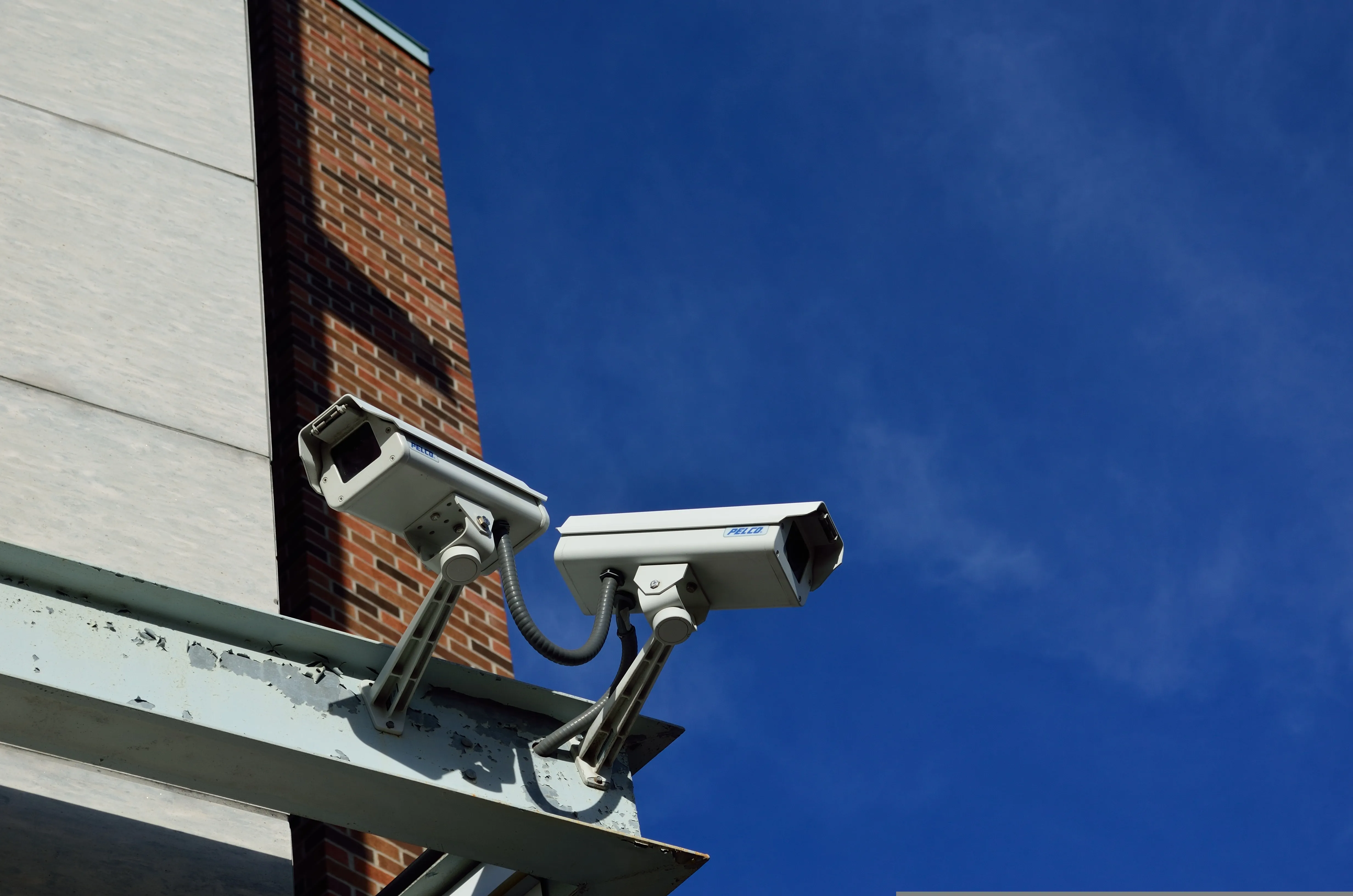 Two white CCTV cameras mounted on a steel bracket on the corner of a brick building against a blue sky
