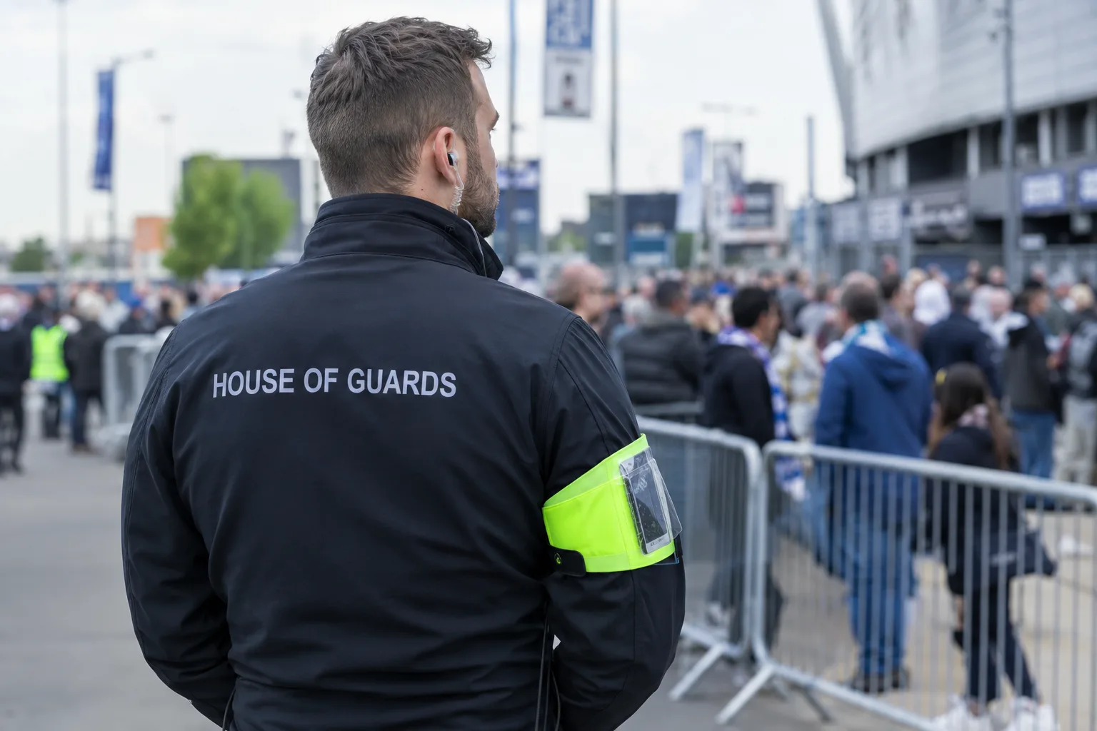 A Harley Garrison officer wearing a hi-vis armband, standing beside a crowd barrier as a large crowd gathers outside a stadium.