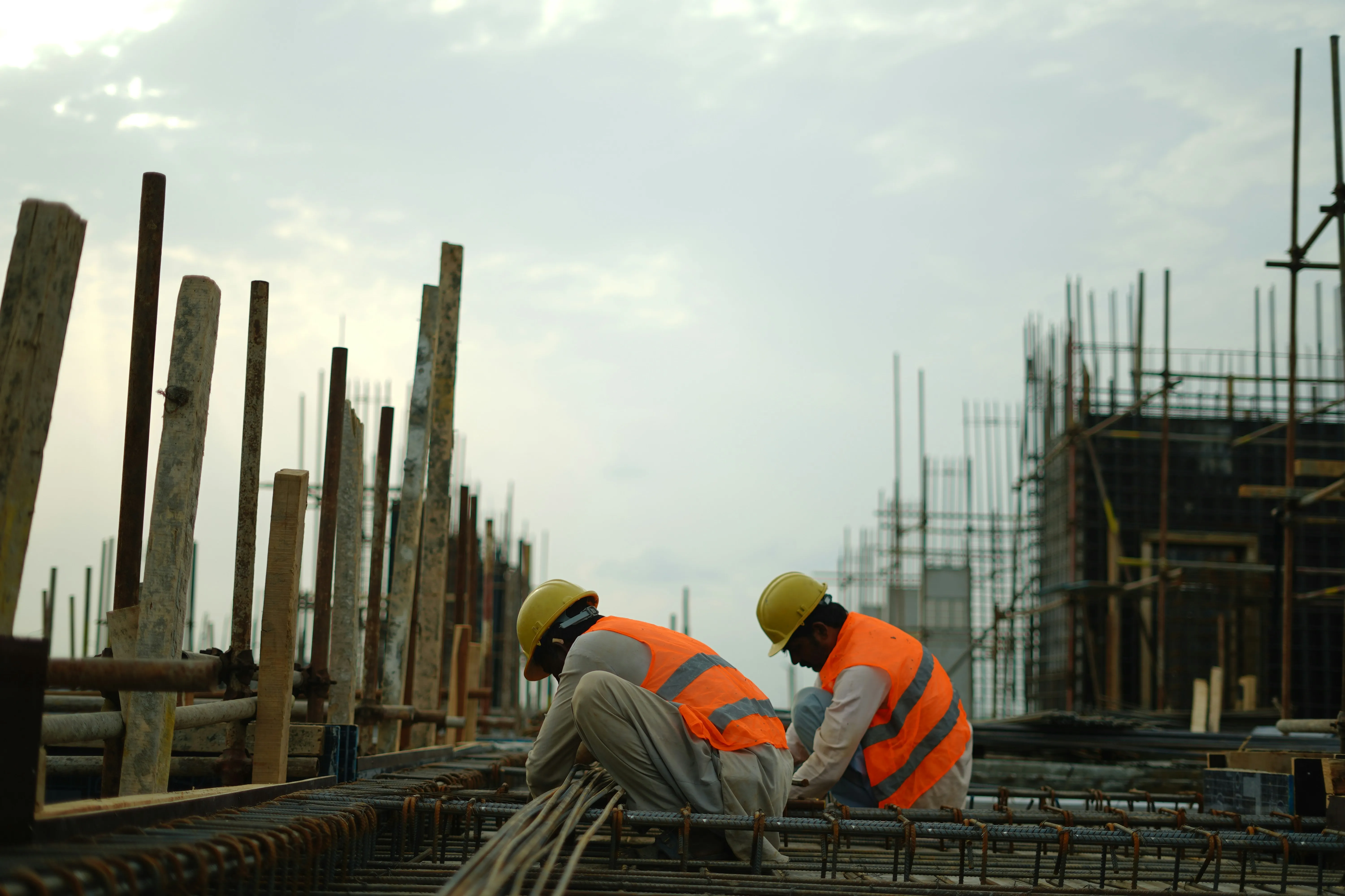 Two construction workers in yellow hard hats and orange high-visibility vests tying steel reinforcement bars on a building site